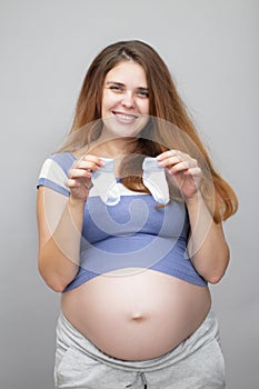 Portrait of happy red-haired pregnant woman with big belly holding little baby socks on isolated grey background