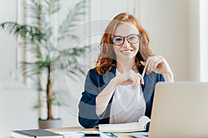 Portrait of happy redhaired woman employee in optical glasses, has satisfied expression, works with modern gadgets, waits for