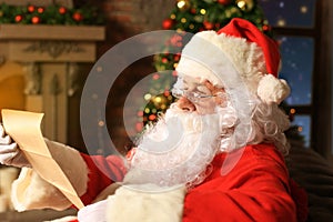 Portrait of happy Santa Claus sitting at his room at home near Christmas tree and reading Christmas letter or wish list.