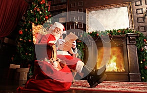 Portrait of happy Santa Claus sitting at his room at home near Christmas tree and reading Christmas letter or wish list.