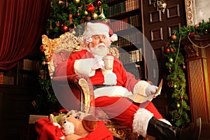 Portrait of happy Santa Claus sitting at his room at home near Christmas tree and reading Christmas letter or wish list.