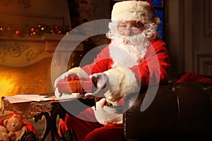 Portrait of happy Santa Claus sitting at his room at home near Christmas tree and reading Christmas letter or wish list.