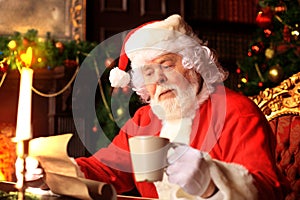 Portrait of happy Santa Claus sitting at his room at home near Christmas tree and reading Christmas letter or wish list.