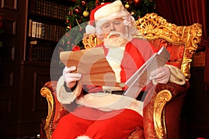 Portrait of happy Santa Claus sitting at his room at home near Christmas tree and reading Christmas letter or wish list.