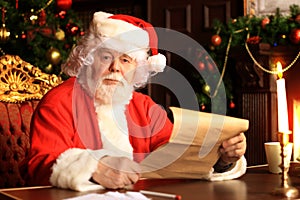 Portrait of happy Santa Claus sitting at his room at home near Christmas tree and reading Christmas letter or wish list.