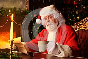 Portrait of happy Santa Claus sitting at his room at home near Christmas tree and reading Christmas letter or wish list.