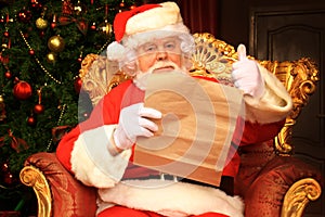 Portrait of happy Santa Claus sitting at his room at home near Christmas tree and reading Christmas letter or wish list.