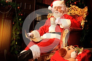 Portrait of happy Santa Claus sitting at his room at home near Christmas tree and reading Christmas letter or wish list.