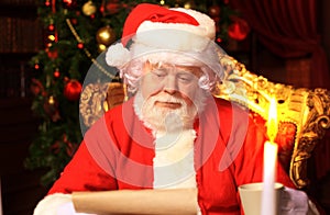 Portrait of happy Santa Claus sitting at his room at home near Christmas tree and reading Christmas letter or wish list.