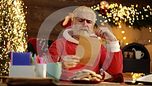 Portrait of happy Santa Claus sitting at his room at home near Christmas tree reading Christmas letter or wish list.