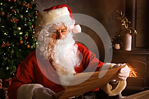 Portrait of happy Santa Claus sitting at his room at home near Christmas tree and reading Christmas letter or wish list