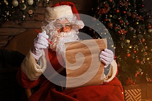 Portrait of happy Santa Claus sitting at his room at home near Christmas tree and reading Christmas letter or wish list