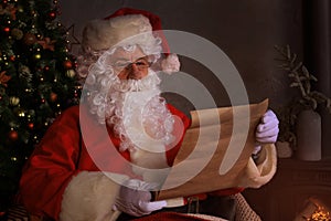 Portrait of happy Santa Claus sitting at his room at home near Christmas tree and reading Christmas letter or wish list