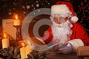 Portrait of happy Santa Claus sitting at his room at home near Christmas tree and reading Christmas letter or wish list