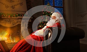 Portrait of happy Santa Claus sitting at his room at home near Christmas tree and reading Christmas letter or wish list.