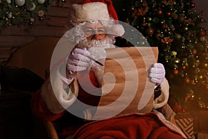 Portrait of happy Santa Claus sitting at his room at home near Christmas tree and reading Christmas letter or wish list