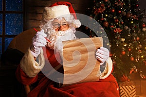 Portrait of happy Santa Claus sitting at his room at home near Christmas tree and reading Christmas letter or wish list