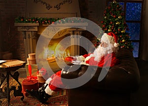 Portrait of happy Santa Claus sitting at his room at home near Christmas tree and reading Christmas letter or wish list.