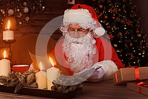 Portrait of happy Santa Claus sitting at his room at home near Christmas tree and reading Christmas letter or wish list