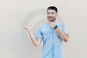 Portrait of happy satisfied handsome young bearded man in blue shirt standing, holding, pointing something and looking at camera