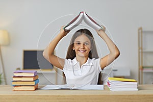 Portrait of happy school girl sitting at desk in classroom and holding book above her head