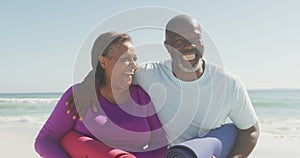 Portrait of happy senior african american couple holding yoga mats on sunny beach