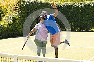 Portrait of happy senior african american couple with rackets embracing on sunny grass tennis court