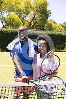 Portrait of happy senior african american couple with tennis rackets embracing on sunny grass court