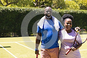 Portrait of happy senior african american couple with tennis rackets embracing on sunny grass court