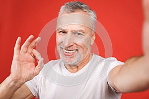 Portrait of happy senior bearded man showing ok sign and taking selfie on smartphone, isolated against red background