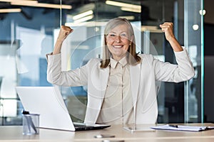 Portrait of a happy senior business woman working in the office using a laptop and rejoicing in success and achievement