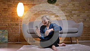 Portrait of happy senior caucasian lady sitting on floor and talking in videochat using smartphone in cozy home