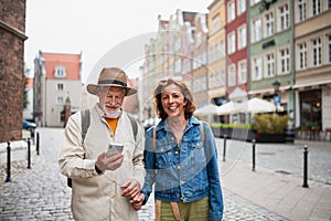 Portrait of happy senior couple tourists smiling, holding hands, using smartphone outdoors in historic town