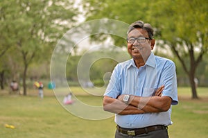 Portrait of a happy senior Indian man wearing a shirt and hands crossed in the outside setting standing and thinking in a park in