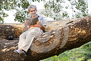portrait happy senior man sitting and reading a book on tree trunk in summer park