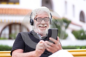 Portrait of happy senior white-haired man with beard sitting in a bench and listening to music smiling. Handsome people using