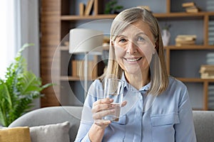 Portrait of happy senior woman in blue shirt looking at camera while raising hand with glass of still water. Positive