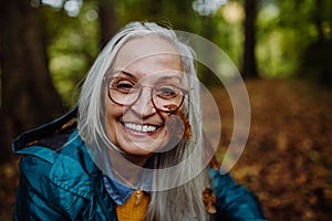 Portrait of happy senior woman with leaf on her cheek on walk outdoors in forest in autumn.