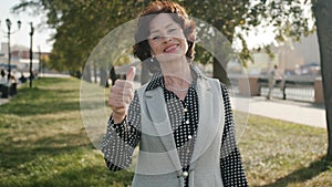 Portrait of happy senior woman smiling and stretching arms with thumbs-up hand gesture outdoors