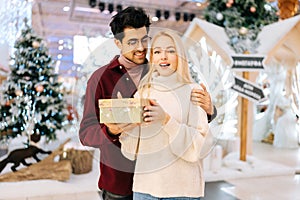 Portrait of happy shocked young girlfriend receiving gift box from loving man standing in hall of celebrate shopping
