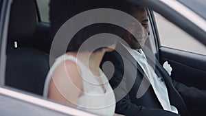Portrait of happy smiling African American groom in elegant suit sitting on driver's seat talking with excited bride in