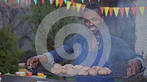 Portrait of happy smiling African American man posing with burger patties on bbq grid outdoors. Joyful handsome young