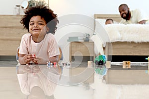 Portrait of happy smiling African boy with black curly hair lying on floor, playing toy in bedroom. Child with reflections on