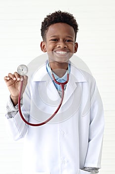 Portrait of happy smiling African boy in lab coat showing stethoscope on white wall room, cute kid pretending to be doctor. Little