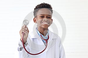 Portrait of happy smiling African boy in lab coat showing stethoscope on white wall room, cute kid pretending to be doctor. Little