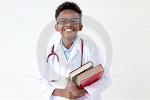 Portrait of happy smiling African boy in lab coat with stethoscope wearing glasses, holding pile of books on white wall room, cute