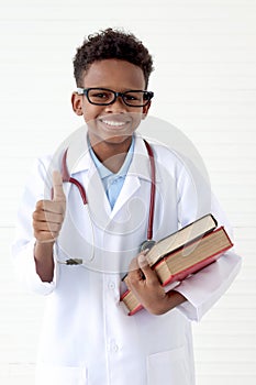 Portrait of happy smiling African boy in lab coat with stethoscope wearing glasses, holding pile of books on white wall room, cute