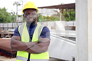 Portrait of happy smiling African engineer worker with safety vest and helmet standing with arms crossed at construction site.