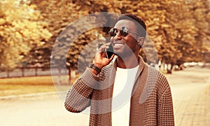 Portrait of happy smiling african man calling on smartphone wearing brown knitted cardigan and sunglasses on city street on autumn