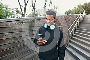 Portrait happy smiling african man with phone, headphones listening to music wearing black hoodie, walking on city